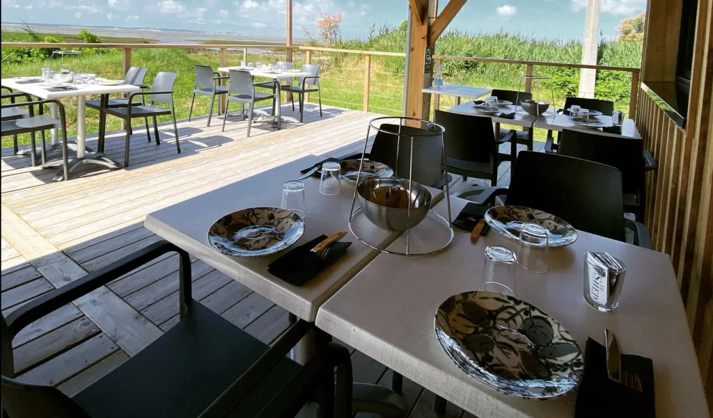 Terrasse en bois d'un restaurant avec vue panoramique sur l'estuaire et les marais, tables dressées pour dégustation de produits locaux et vins de Bordeaux