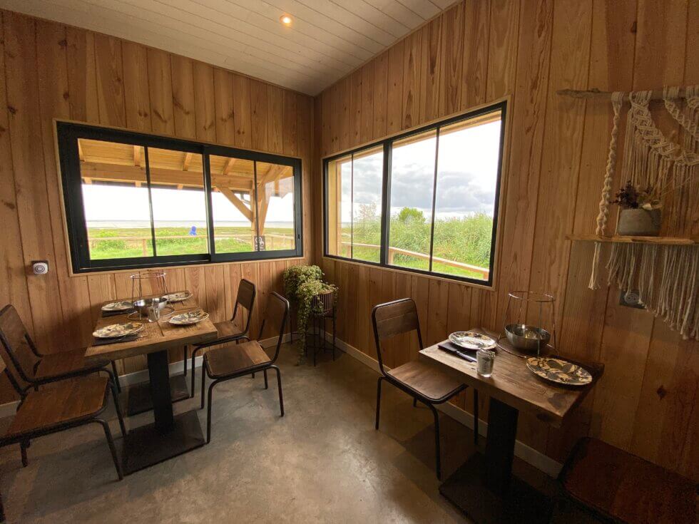 Salle à manger intérieure du Shed, avec des tables en bois individuelles dressées, des chaises en métal et bois, et des vues sur le paysage extérieur via les fenêtres, ambiance rustique et naturelle